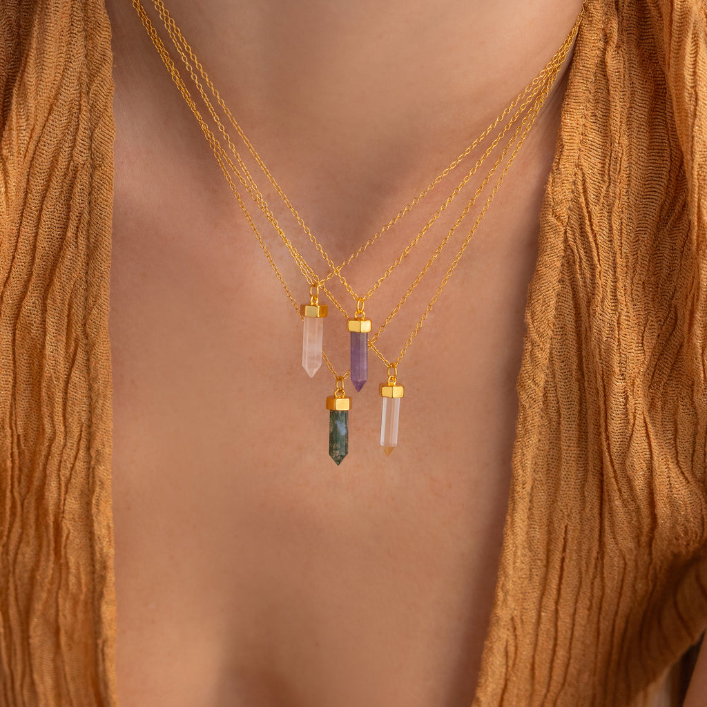 Close-up of three layered gold Clear Quartz Necklaces featuring colorful crystal pendants, including a hexagonal point pendant, styled over a brown textured top—ideal for crystal jewelry lovers.