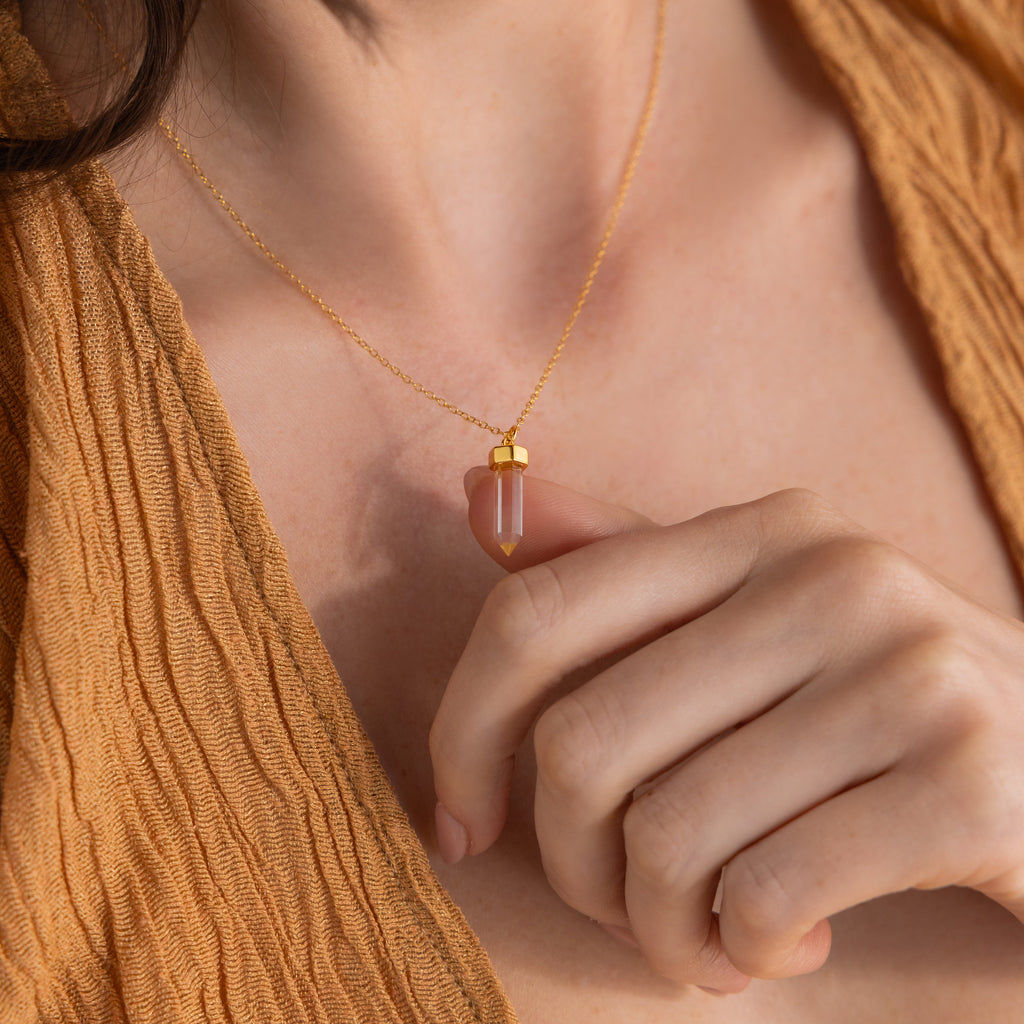 A woman wears the Clear Quartz Necklace featuring a gold hexagonal point pendant, gently holding it in her hand and pairing it with a brown top.