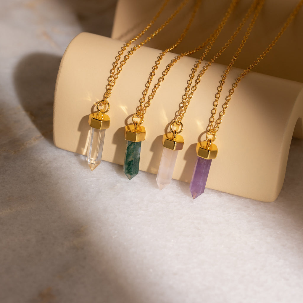 The Clear Quartz Necklace features a gold chain with a hexagonal point pendant, displayed elegantly on a beige holder alongside variations in green, pink, and purple crystal jewelry.