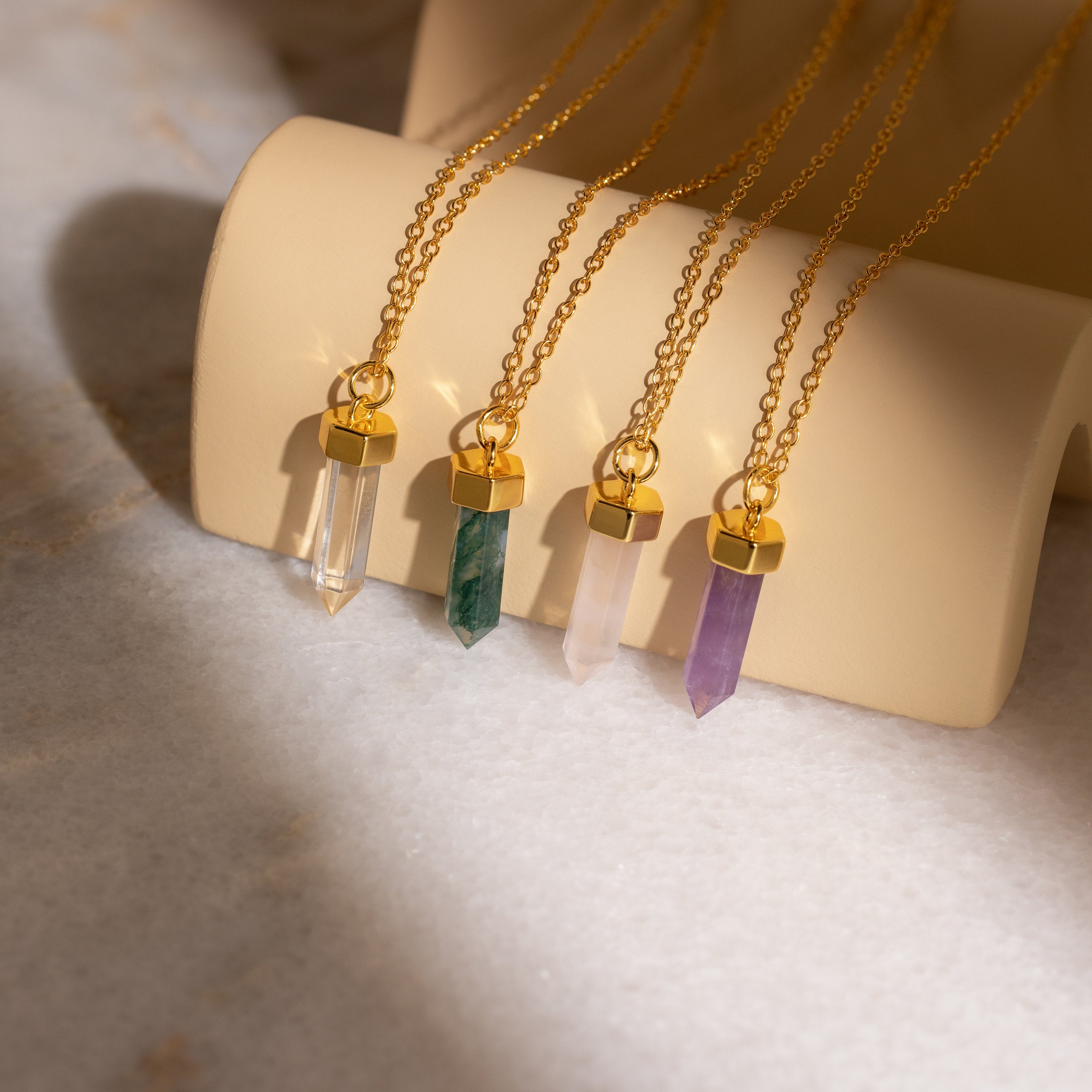 The Clear Quartz Necklace features a gold chain with a hexagonal point pendant, displayed elegantly on a beige holder alongside variations in green, pink, and purple crystal jewelry.