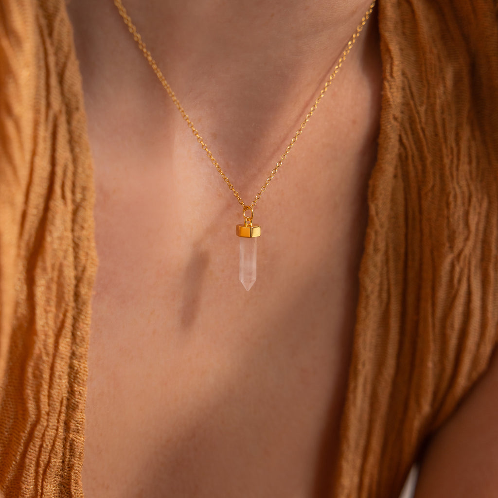 Worn by someone in a brown textured top, the Crystal Quartz Necklaces feature a gold chain and clear hexagonal point pendant that radiates positive energy.