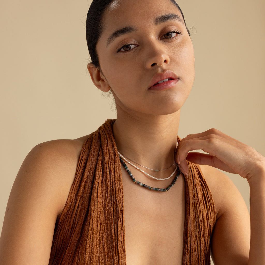 A woman in a brown textured top wears the Moss Agate Beaded Necklace layered with others, gazing at the camera against a beige backdrop—an effortless showcase of earthy elegance.