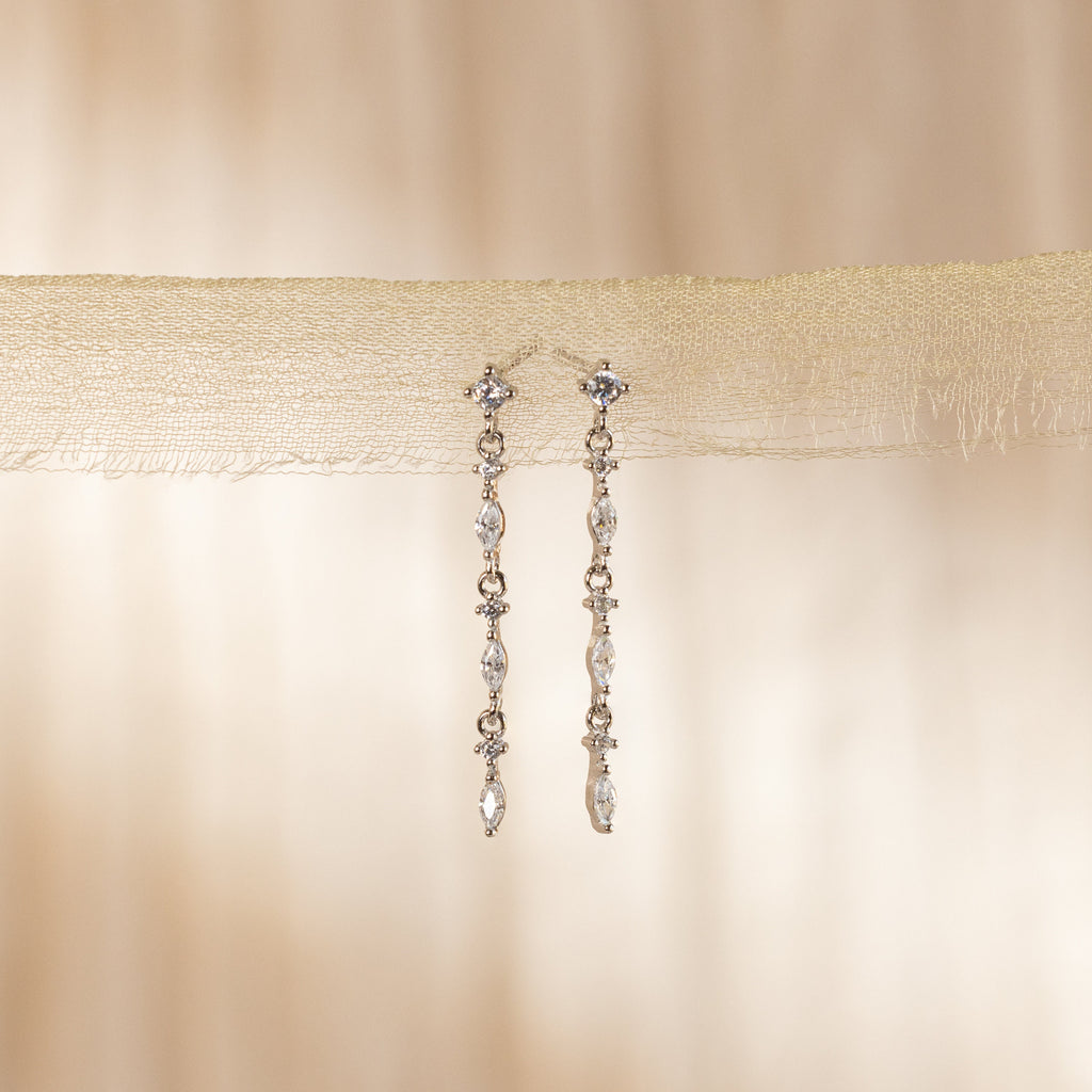 The Dainty Diamond Drop Earrings feature sparkling stones dangling from sheer beige fabric, ideal as wedding or bridesmaid jewelry for an elegant touch against a soft background.