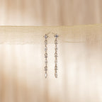 The Dainty Diamond Drop Earrings feature sparkling stones dangling from sheer beige fabric, ideal as wedding or bridesmaid jewelry for an elegant touch against a soft background.