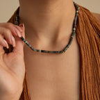 Wearing a brown textured top, a person showcases earthy elegance while holding the Moss Agate Beaded Necklace, whose mixed color beads add natural charm to their style.