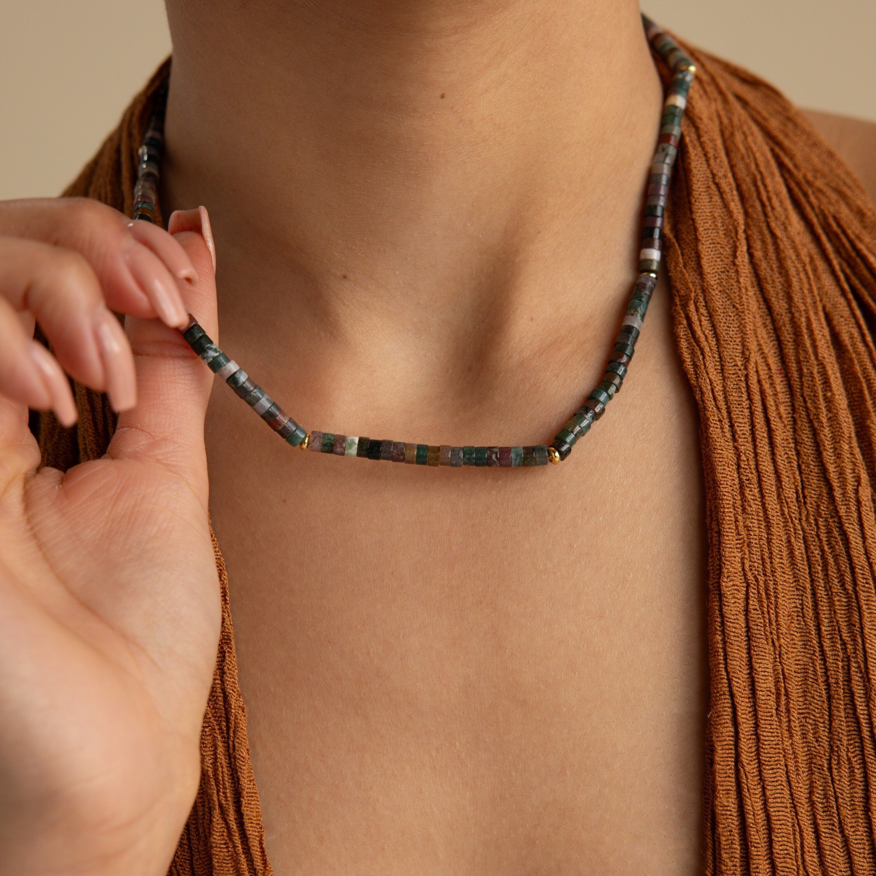 Wearing a brown textured top, a person showcases earthy elegance while holding the Moss Agate Beaded Necklace, whose mixed color beads add natural charm to their style.