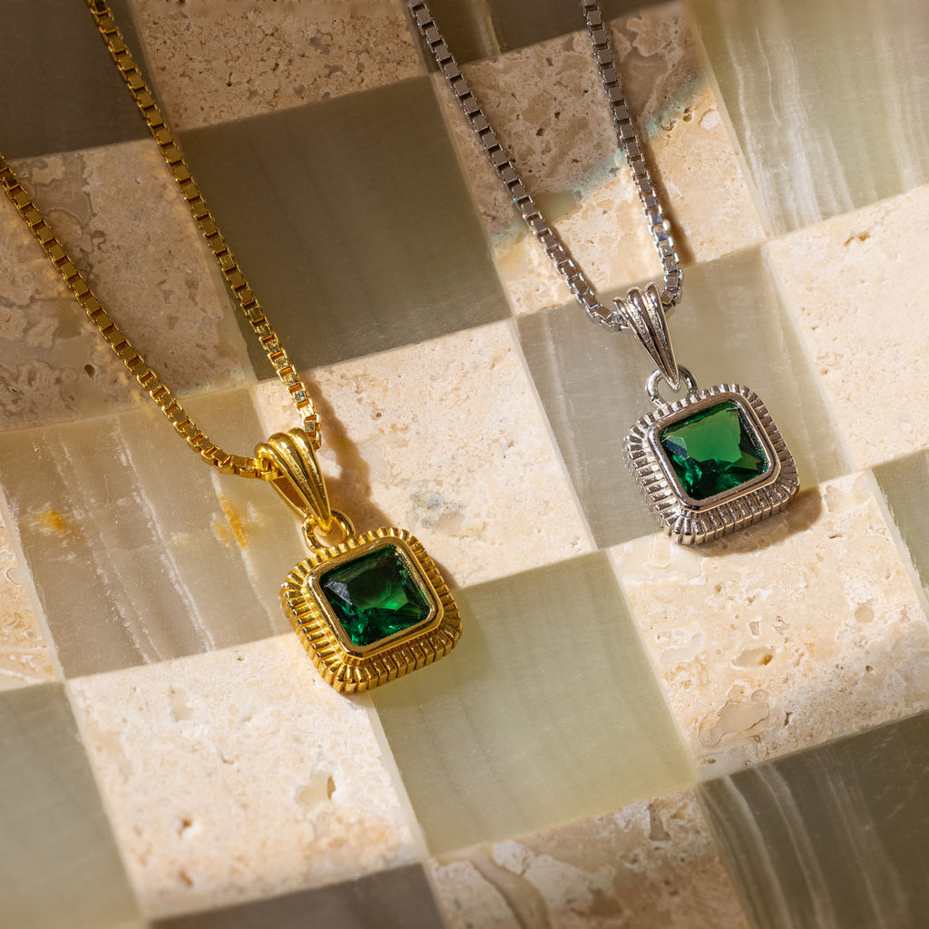 Two Vintage Emerald Pendant Necklaces—one gold, one silver—are displayed on a beige and green checkered surface, perfect for adding an Art Deco touch to your look.