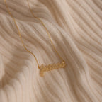 The Pave Handwriting Necklace, spelling 'Jessica' in gold, rests on a ribbed cream fabric surface—an elegant statement jewelry piece.
