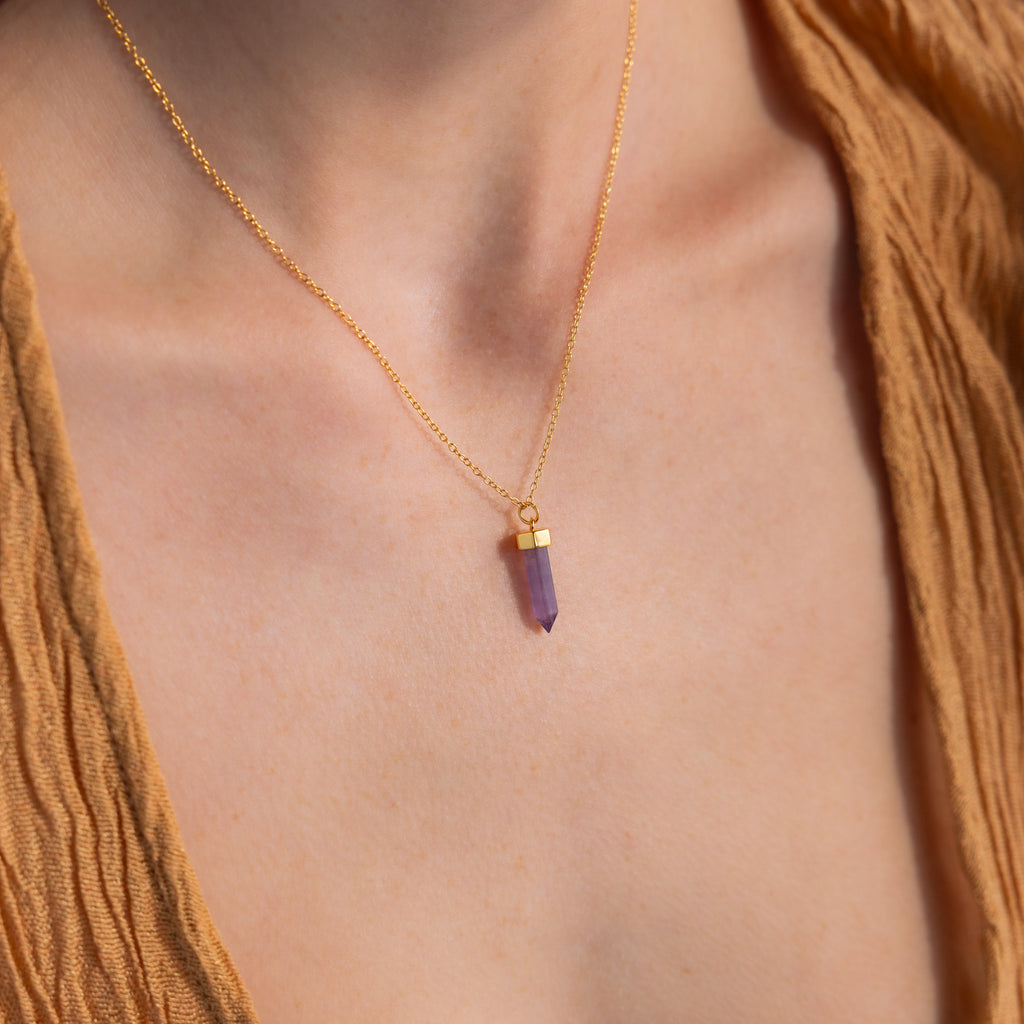 An Amethyst Quartz Necklace with a purple crystal pendant is worn around the neck, styled with a brown textured top—making it an elegant and unique gift for any occasion.