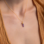 An Amethyst Quartz Necklace with a purple crystal pendant is worn around the neck, styled with a brown textured top—making it an elegant and unique gift for any occasion.