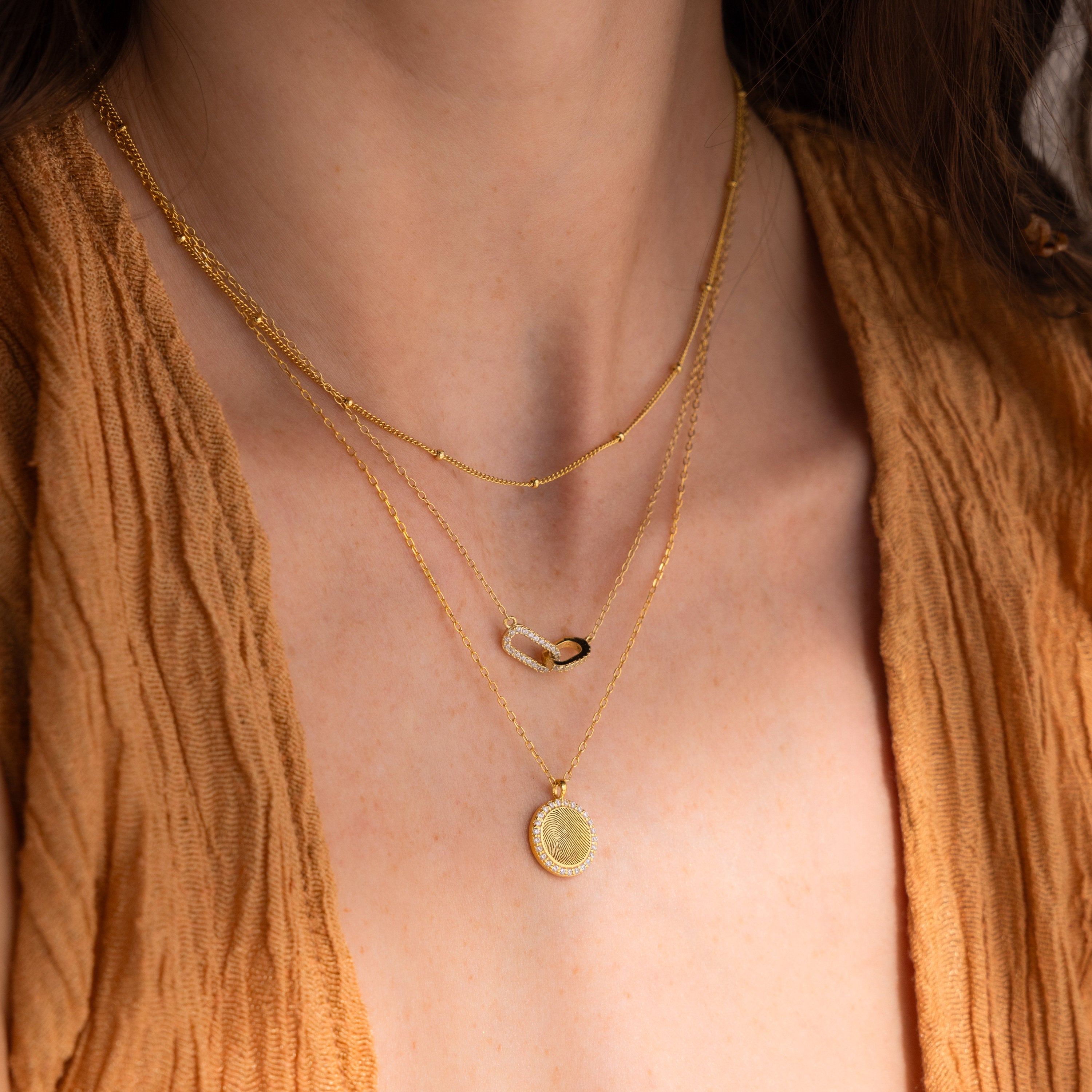 Woman wearing the Pave Fingerprint Coin Necklace, a three-layered gold piece with a custom engraved pendant, styled over a textured tan V-neck top.