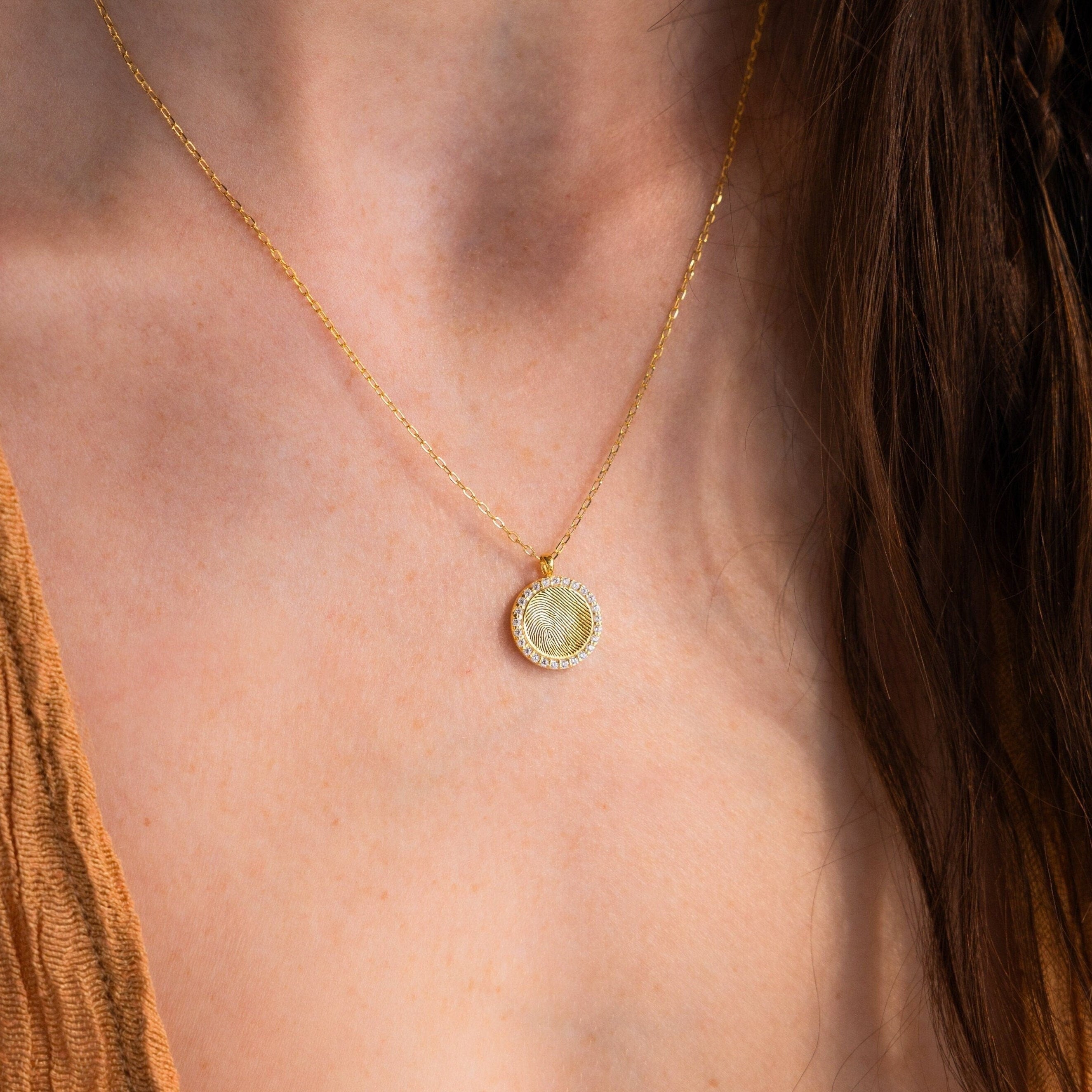 The Pave Fingerprint Coin Necklace features a round, custom-engraved pendant on a gold chain—ideal as a memorial gift or personalized keepsake. Shown resting on the neck above a tan textured top.