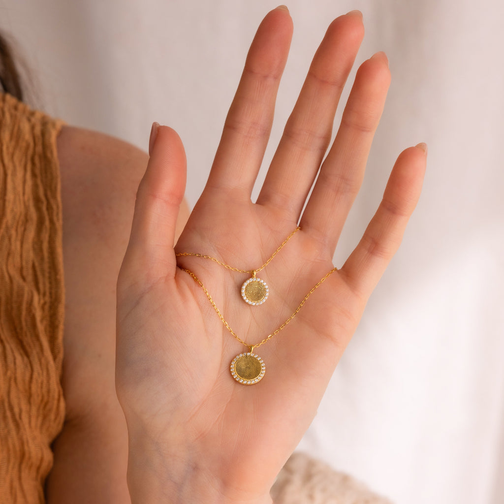 A hand holds two Pave Fingerprint Coin Necklaces against a neutral background—custom-engraved keepsakes, ideal as memorial gifts.