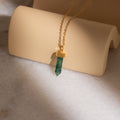 The Agate Quartz Necklace, featuring a marbled green gemstone charm pendant on a gold chain, is displayed on a curved beige stand in soft sunlight.