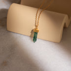 The Crystal Quartz Necklaces feature a gold chain with a hexagonal green crystal pendant, displayed on a curved beige surface to inspire positivity.