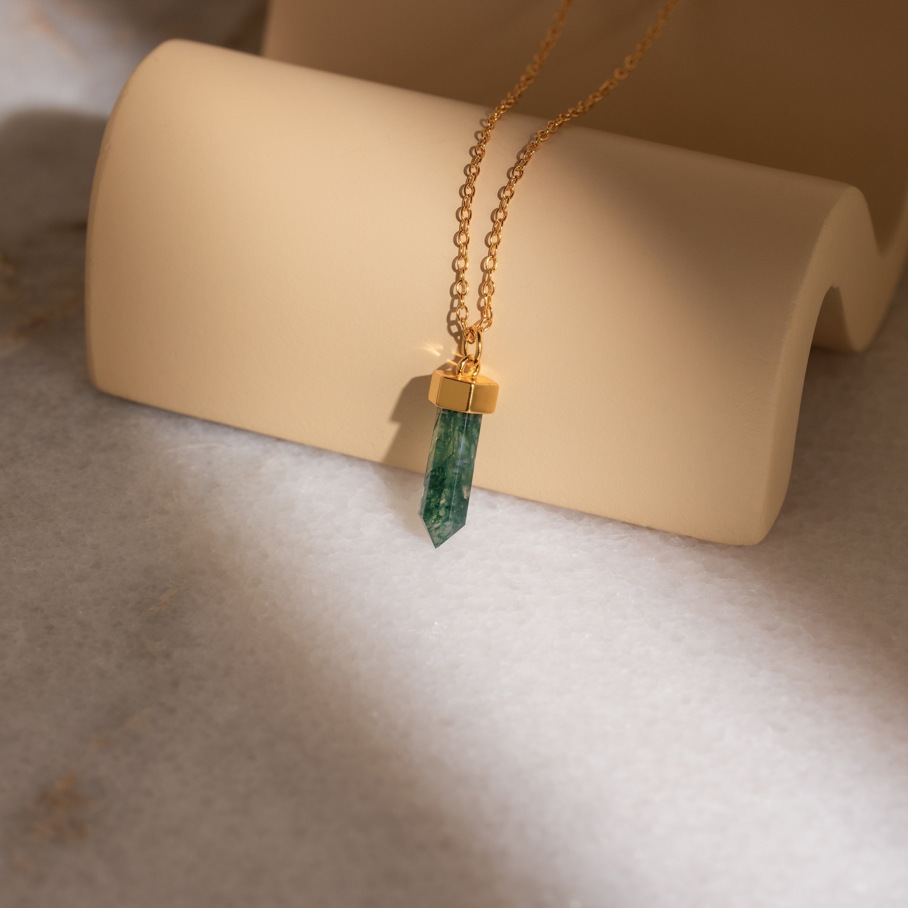 The Crystal Quartz Necklaces feature a gold chain with a hexagonal green crystal pendant, displayed on a curved beige surface to inspire positivity.