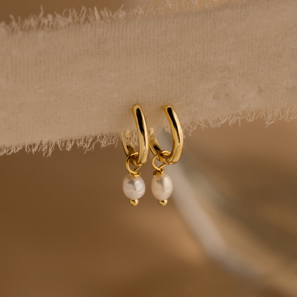 Close-up of Dainty Gold Pearl Huggies hanging from a sheer beige fabric.