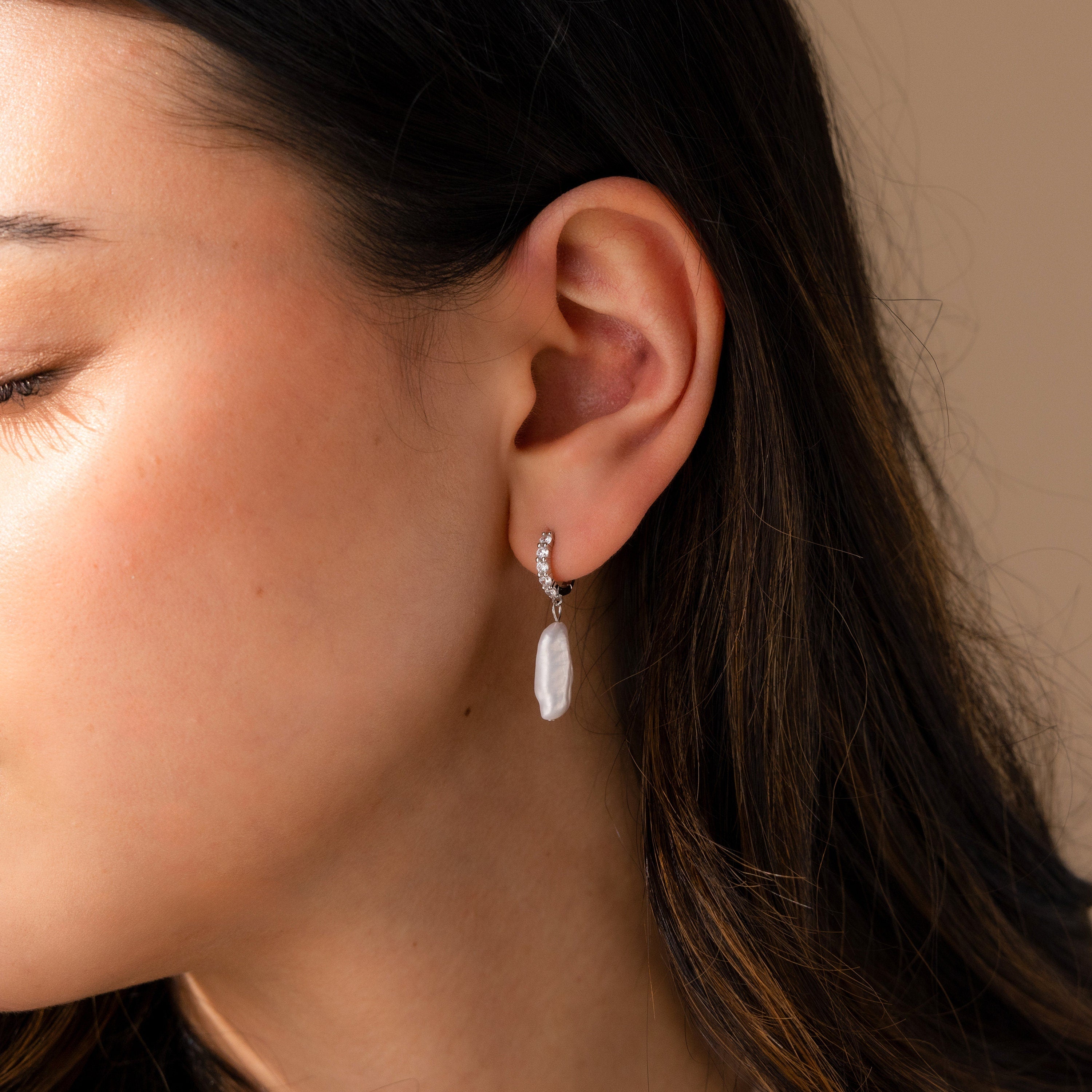 A close-up side view of a woman wearing Baroque Pearl Pave Huggies, featuring a dangling baroque pearl from a silver hoop.
