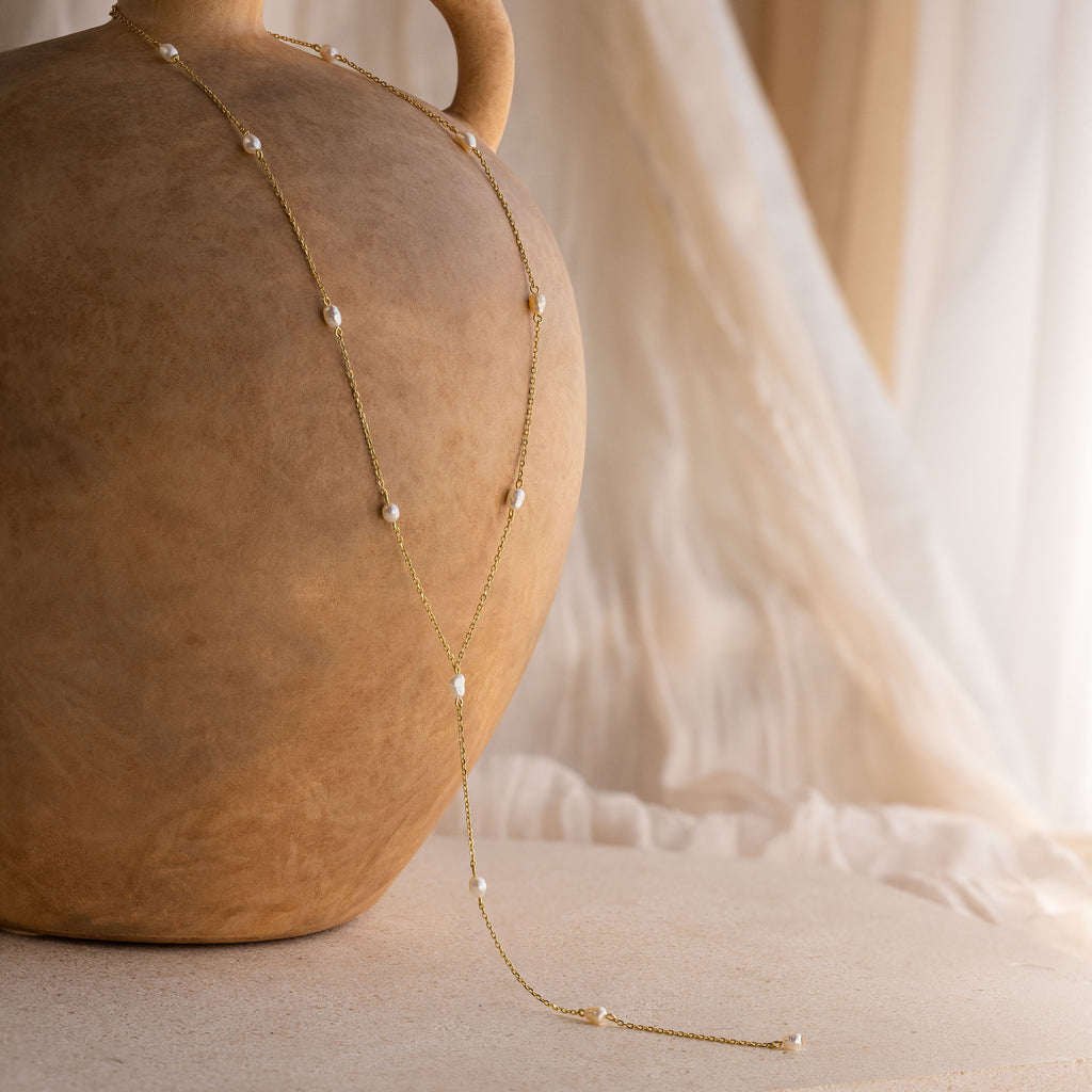 Pearl Station Lariat Necklace adorned with pearl charms draped over a large clay vase, set against a soft neutral background.