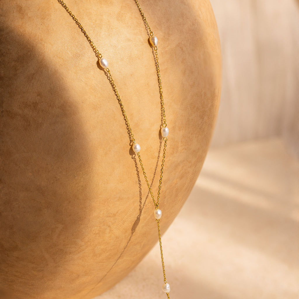 Pearl Station Lariat Necklace featuring small pearl charms elegantly draped over a round, light brown textured surface in soft sunlight.
