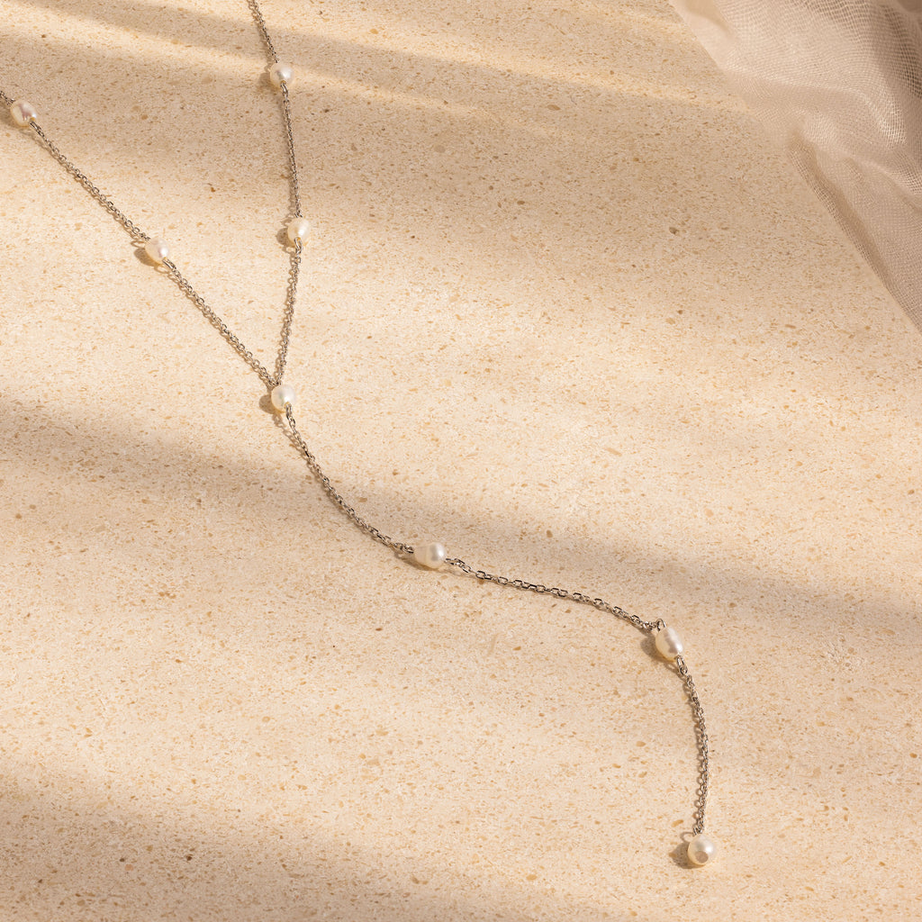 The Pearl Station Lariat Necklace features small pearl charms on a silver chain, displayed on a beige textured surface and illuminated by gentle natural light with soft shadows.