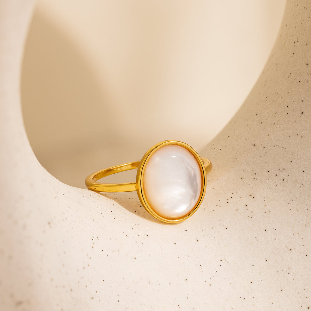 The Mother of Pearl Signet Ring features a vintage-inspired design with a round white pearl set in gold, showcased on a textured beige background.