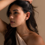 A woman with long brown hair, wearing a beige top and Freshwater Pearl Studs, poses gracefully with one hand on her head, showcasing the timeless elegance of the Freshwater Pearl Studs.
