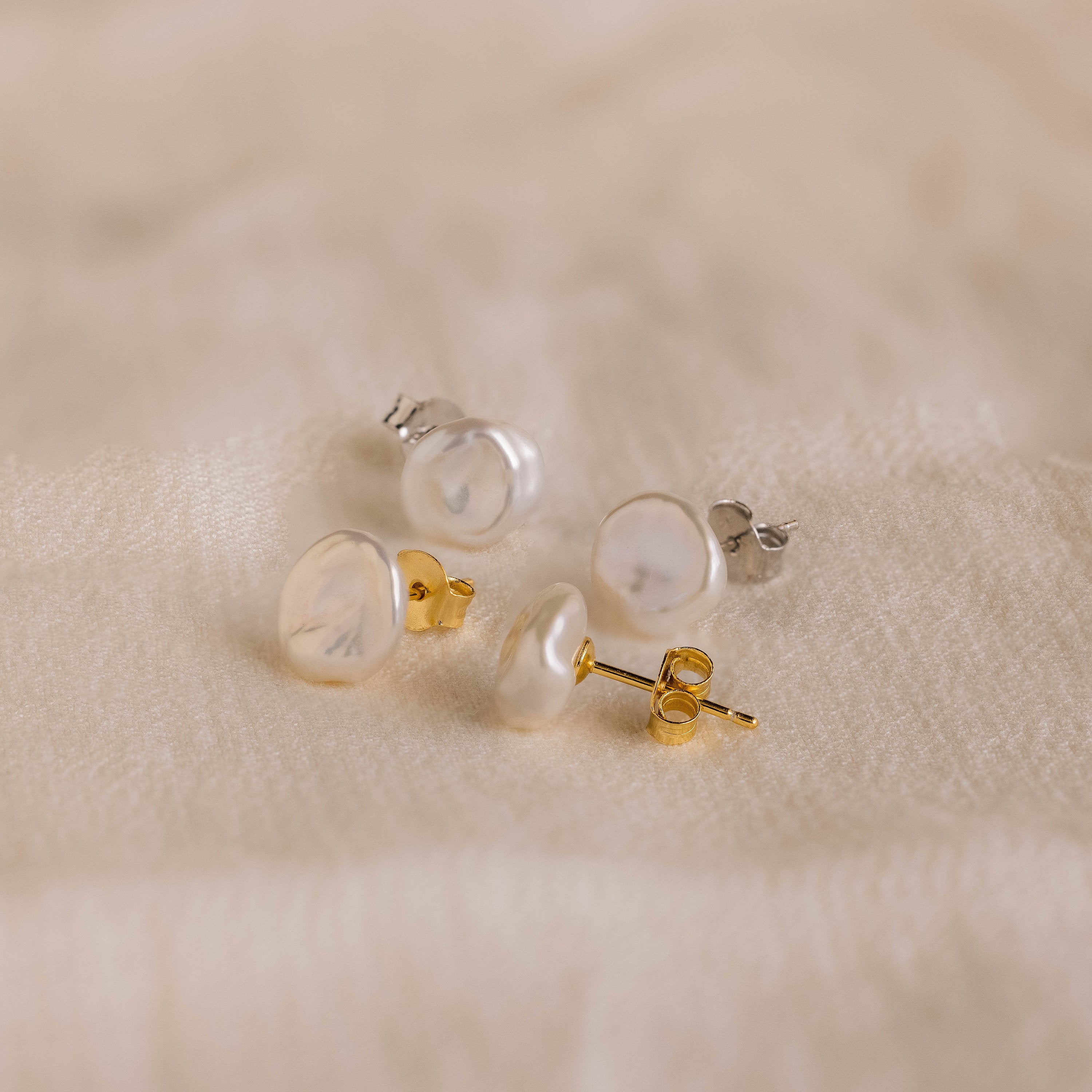 Two pairs of Freshwater Pearl Studs, featuring both gold and silver backs, are displayed on soft beige fabric, showcasing their timeless elegance.