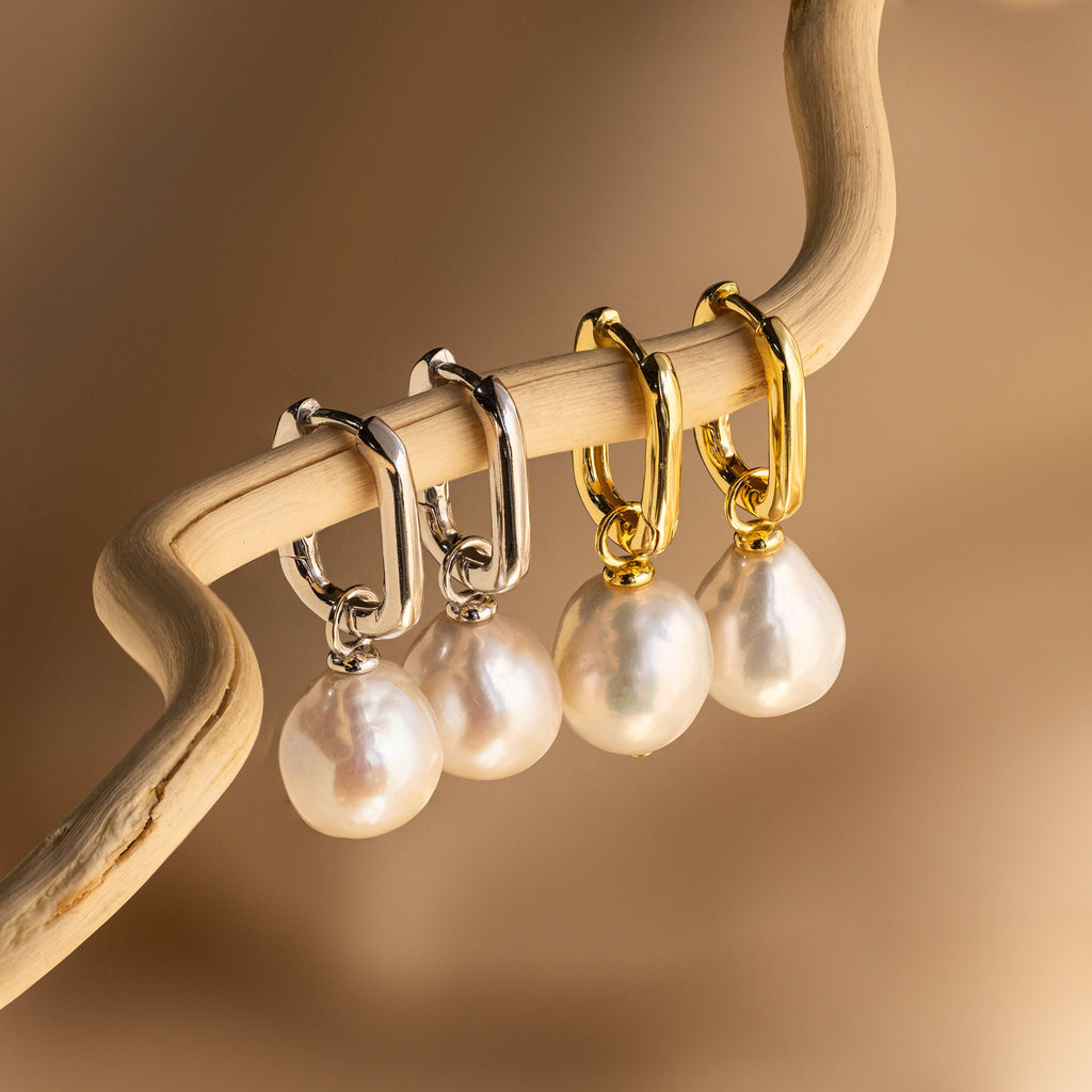 Two pairs of Freshwater Pearl Drop Hoops—one in silver, one in gold—elegantly hang from a curved beige branch, highlighting their timeless style.