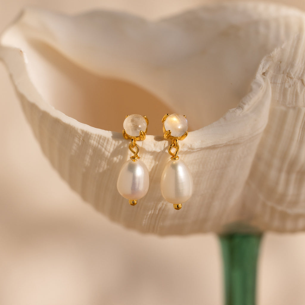 The Moonstone Pearl Drop Earrings from our collection are displayed on a white, shell-shaped surface with a soft background.