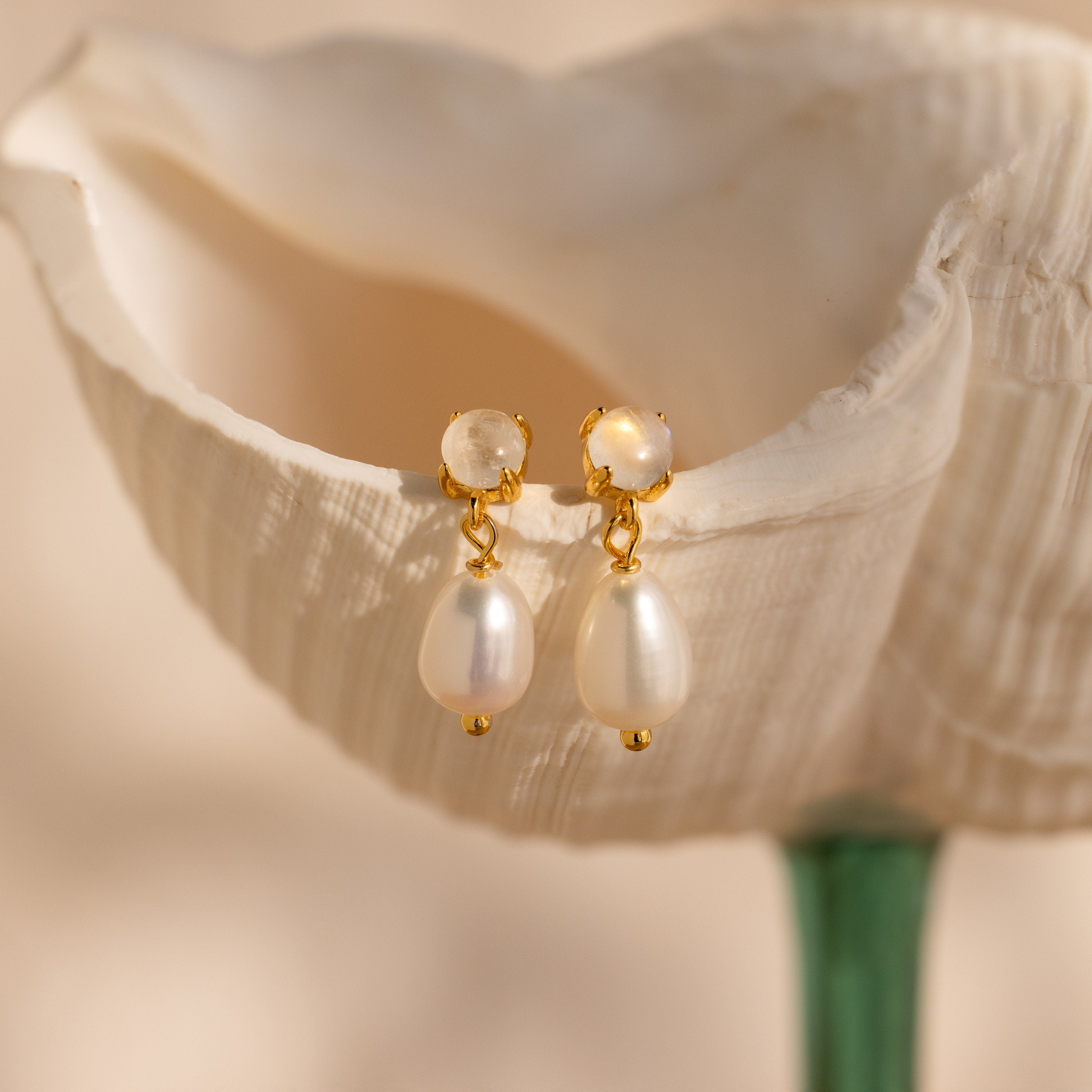 The Moonstone Pearl Drop Earrings from our collection are displayed on a white, shell-shaped surface with a soft background.