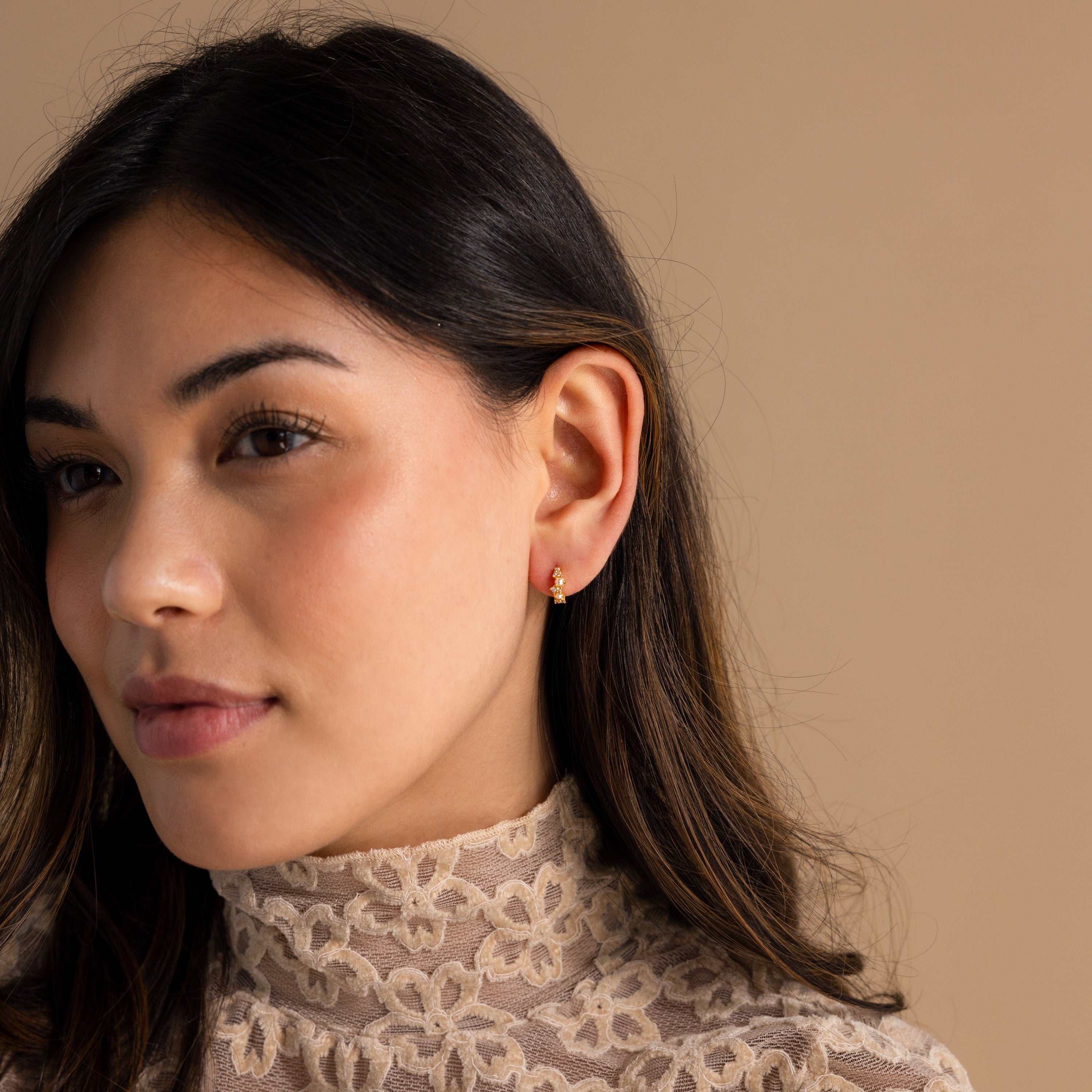 Woman with long dark hair wears a lace top and Pearl Diamond Cluster Huggies, set against a beige background.