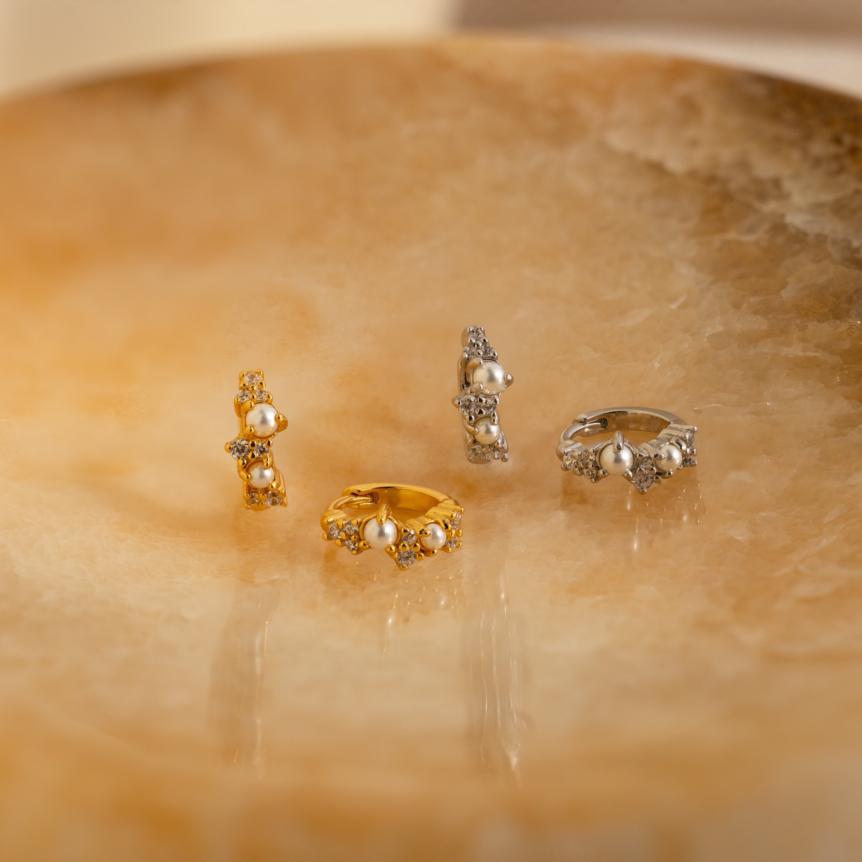 Two gold and two silver Pearl Diamond Cluster Huggies, radiant like luxurious pearls, are elegantly displayed on a polished, marble-like beige surface.