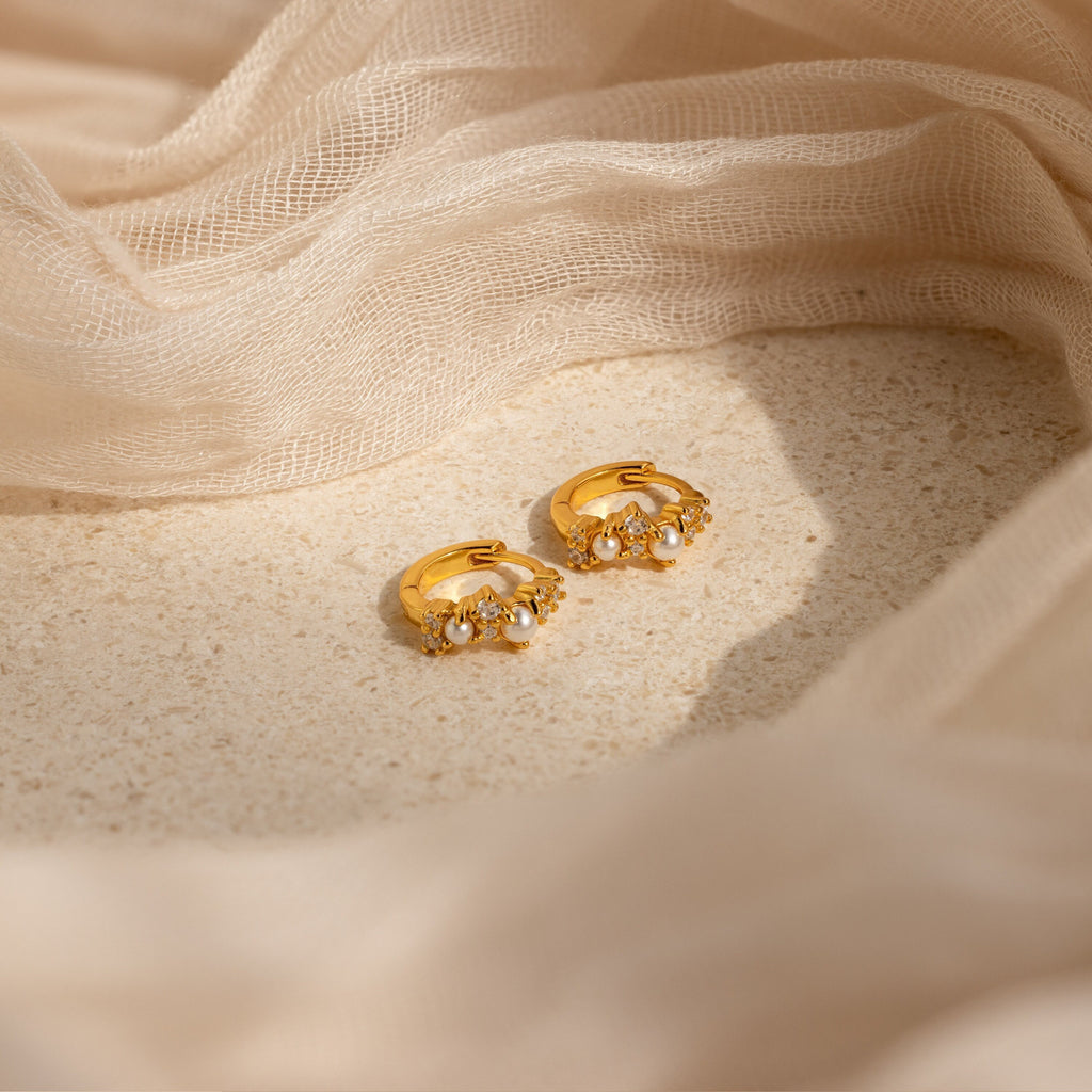 The Pearl Diamond Cluster Huggies, featuring petite pearls and sparkling diamonds, rest elegantly on a textured surface draped with soft beige fabric.