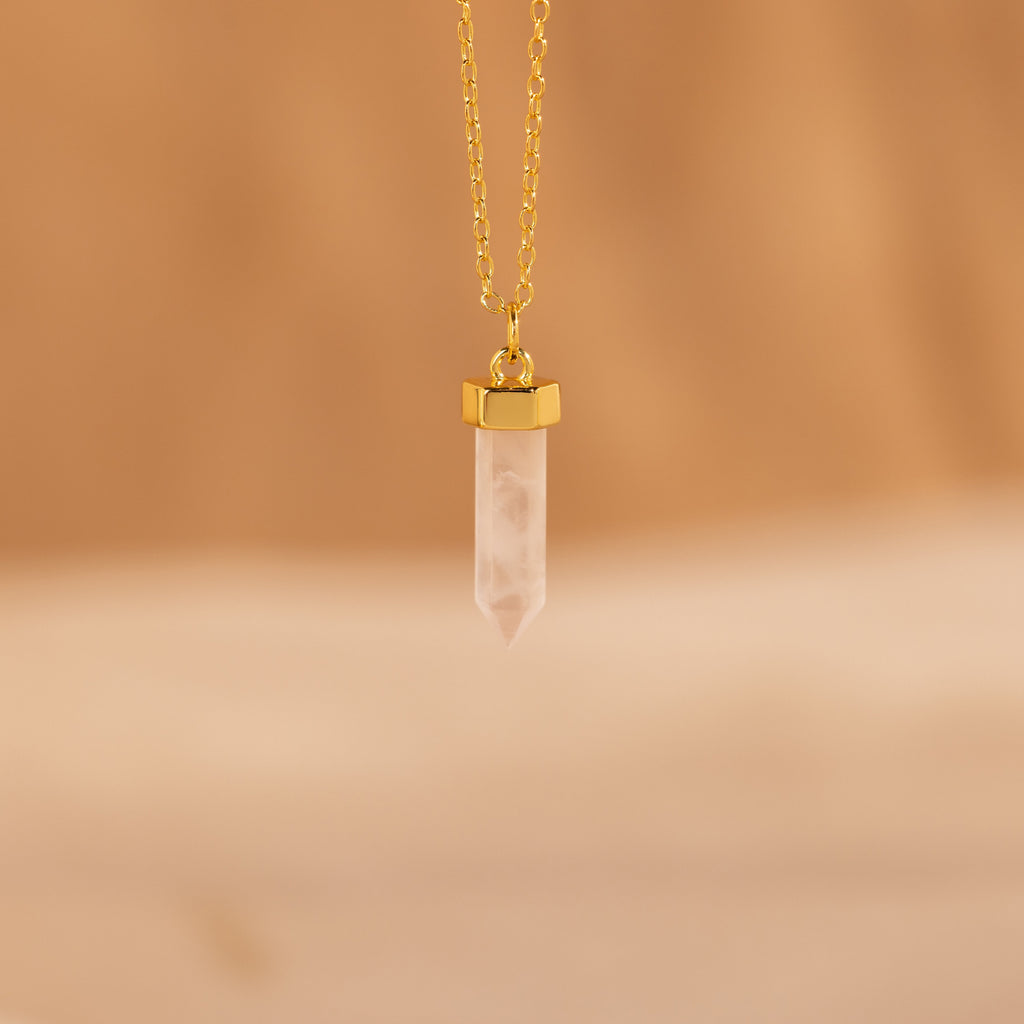 The Rose Quartz Necklace, featuring a gold chain and pointed crystal pendant known as the stone of love, is elegantly displayed against a soft beige background.