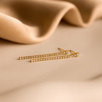 Gold and diamond earrings rest on a beige surface with soft fabric in the background, highlighting the elegance of Diamond Tennis Dangle Earrings.