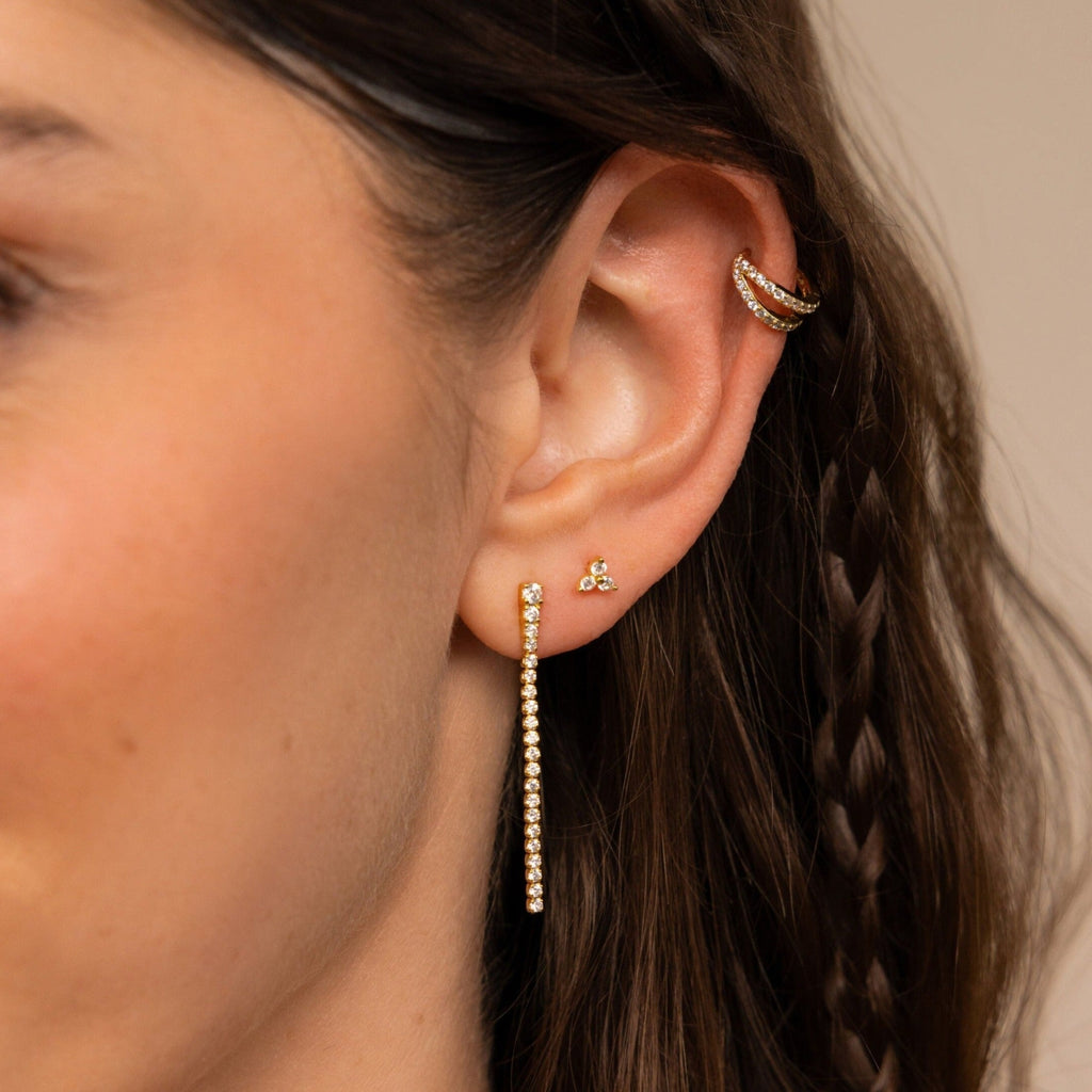 Close-up of a woman's ear with three gold earrings, featuring Diamond Tennis Dangle Earrings, and a small braid in her brown hair.