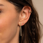 Close-up of a woman's ear with three gold earrings, featuring Diamond Tennis Dangle Earrings, and a small braid in her brown hair.