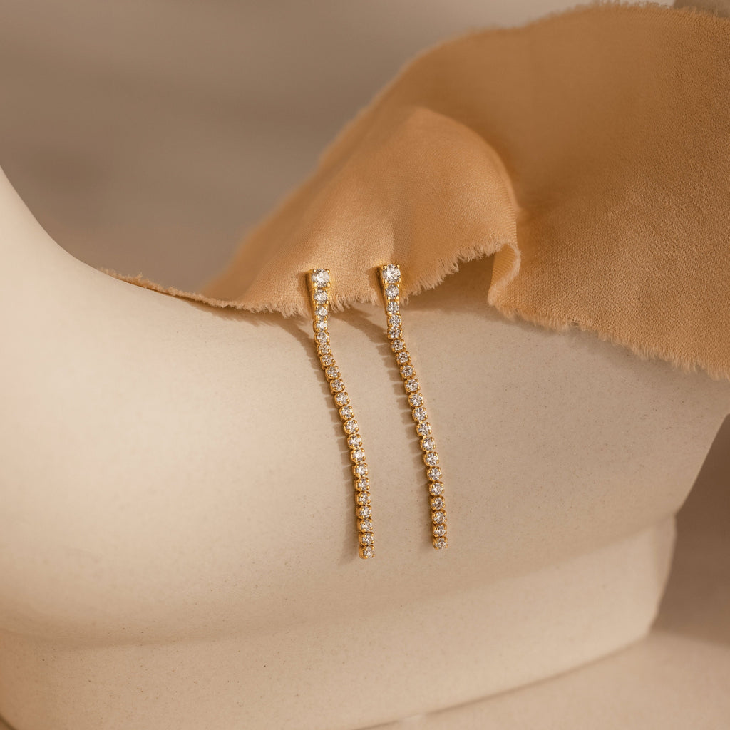 Diamond Tennis Dangle Earrings, featuring rows of sparkling diamonds, are shown on a cream-colored mannequin draped in tan fabric.