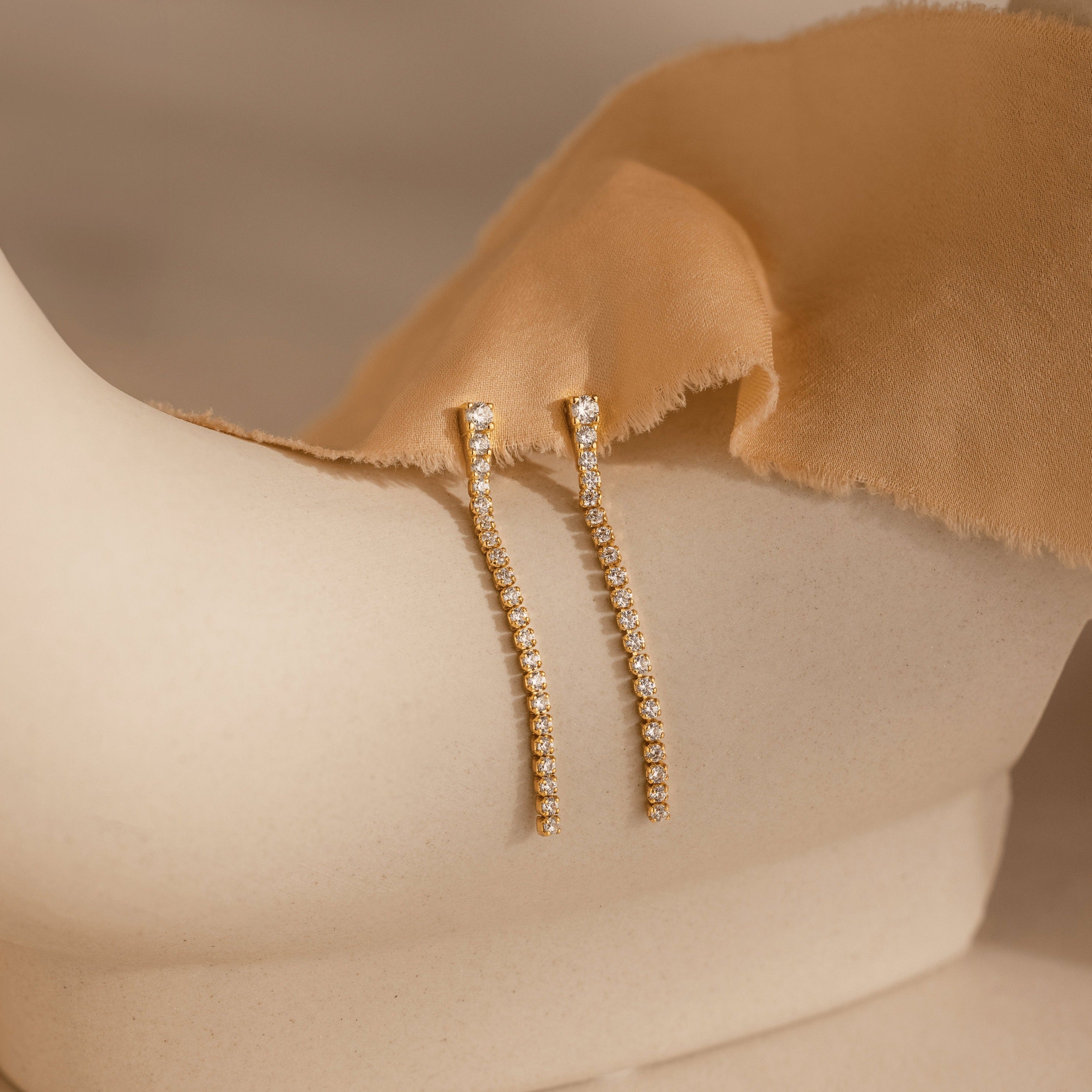 Diamond Tennis Dangle Earrings, featuring rows of sparkling diamonds, are shown on a cream-colored mannequin draped in tan fabric.