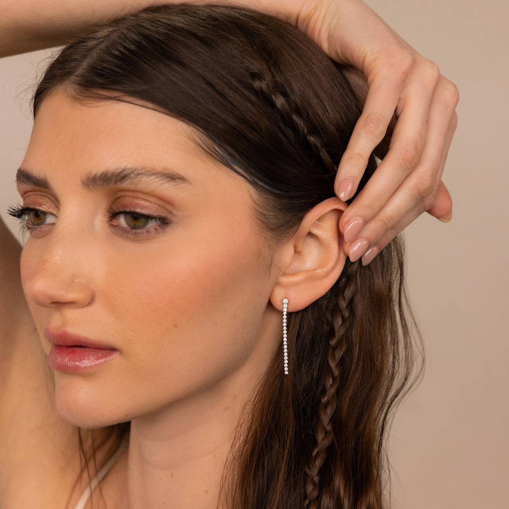 A woman with long brown braided hair touches her head while wearing elegant Diamond Tennis Dangle Earrings.