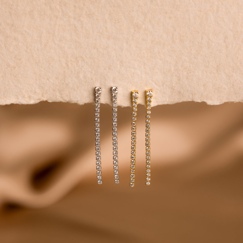 Two pairs of Diamond Tennis Dangle Earrings—one in silver, one in gold—shine on a textured beige surface, highlighting their elegant design that’s perfect for any occasion.