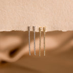 Two pairs of Diamond Tennis Dangle Earrings—one in silver, one in gold—shine on a textured beige surface, highlighting their elegant design that’s perfect for any occasion.