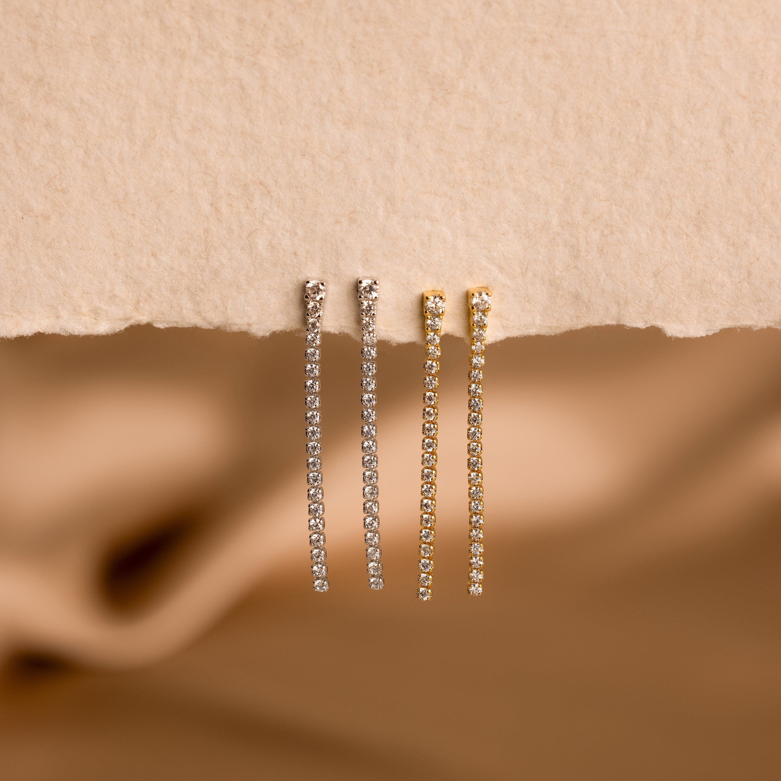 Two pairs of Diamond Tennis Dangle Earrings—one in silver, one in gold—shine on a textured beige surface, highlighting their elegant design that’s perfect for any occasion.