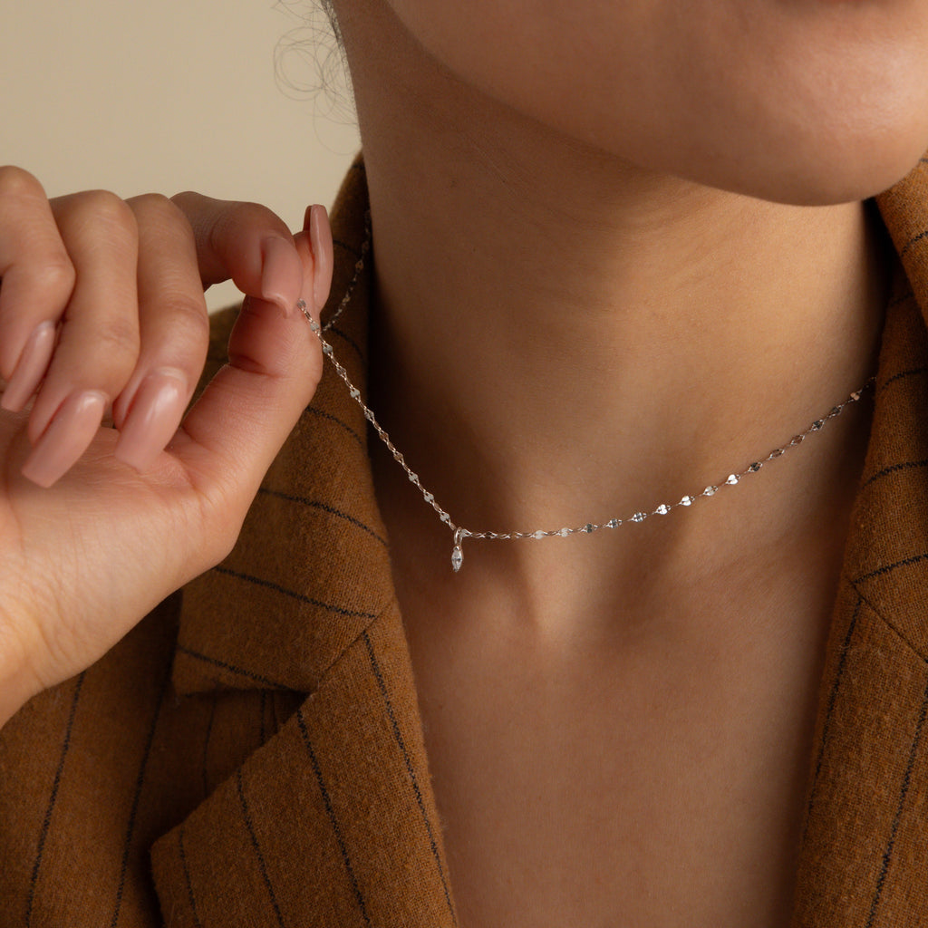 A person in a brown blazer holds the Dainty Marquise Pendant Necklace, featuring a delicate mirror twist chain and small pendant, near their neck.