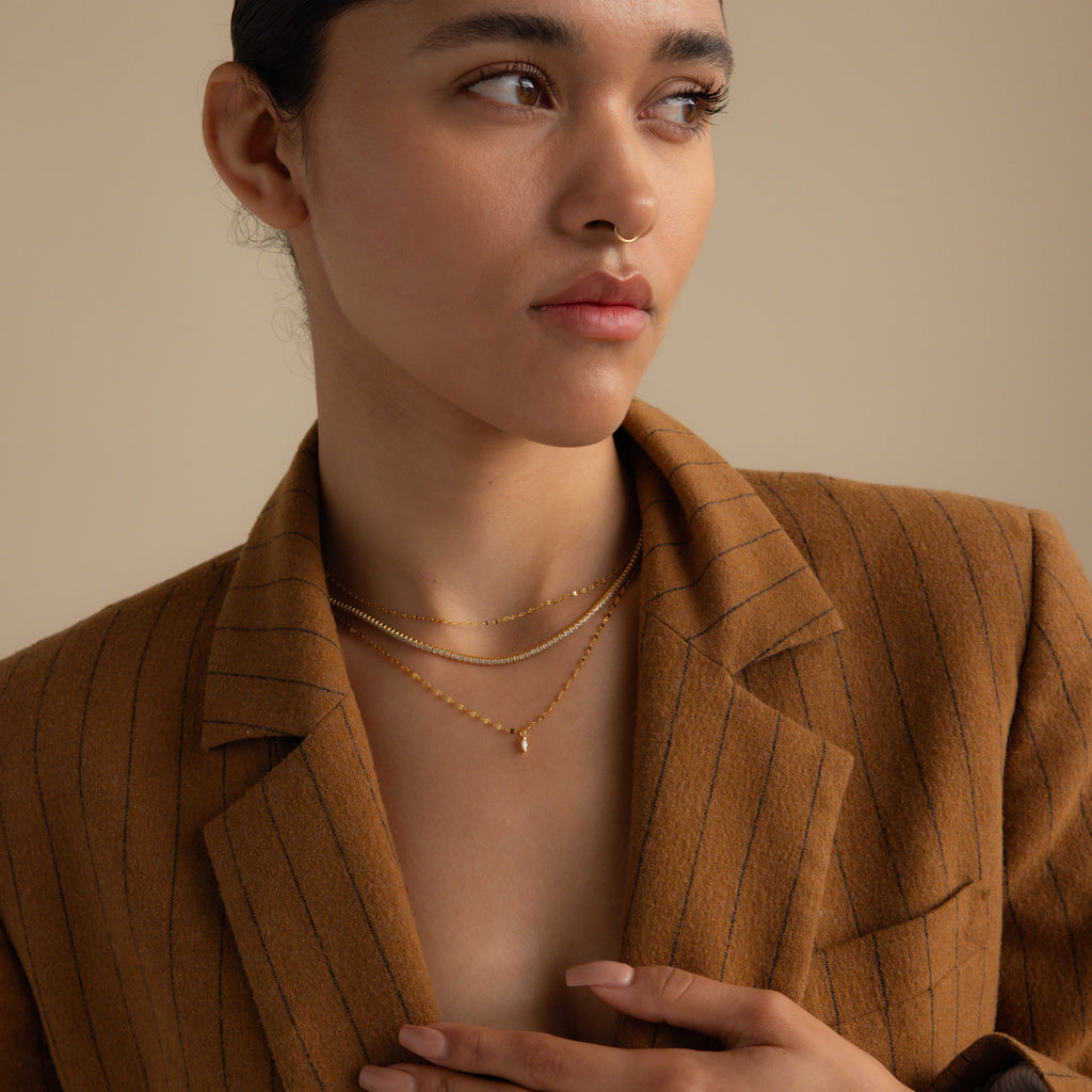 Woman in a brown pinstripe blazer, wearing layered gold necklaces including the Dainty Marquise Pendant Necklace and a mirror twist chain, looking to the side against a neutral background.