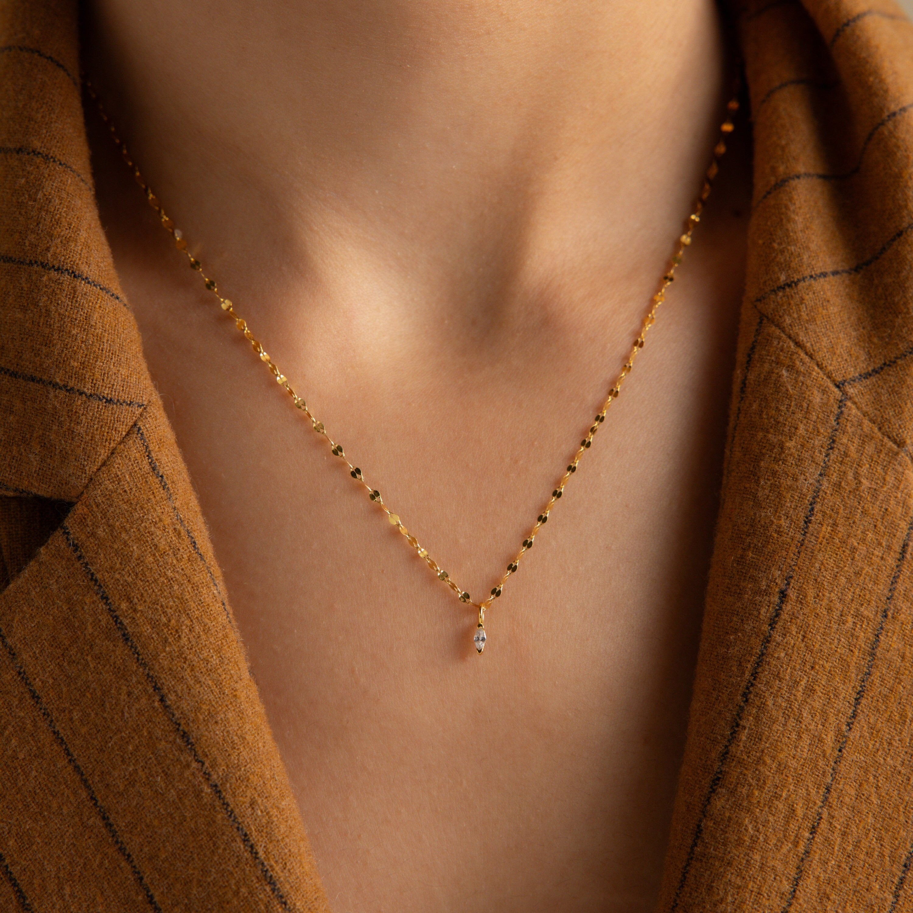 The Dainty Marquise Pendant Necklace, featuring a small gold marquise pendant and mirror twist chain, rests elegantly on someone in a brown pinstripe jacket.