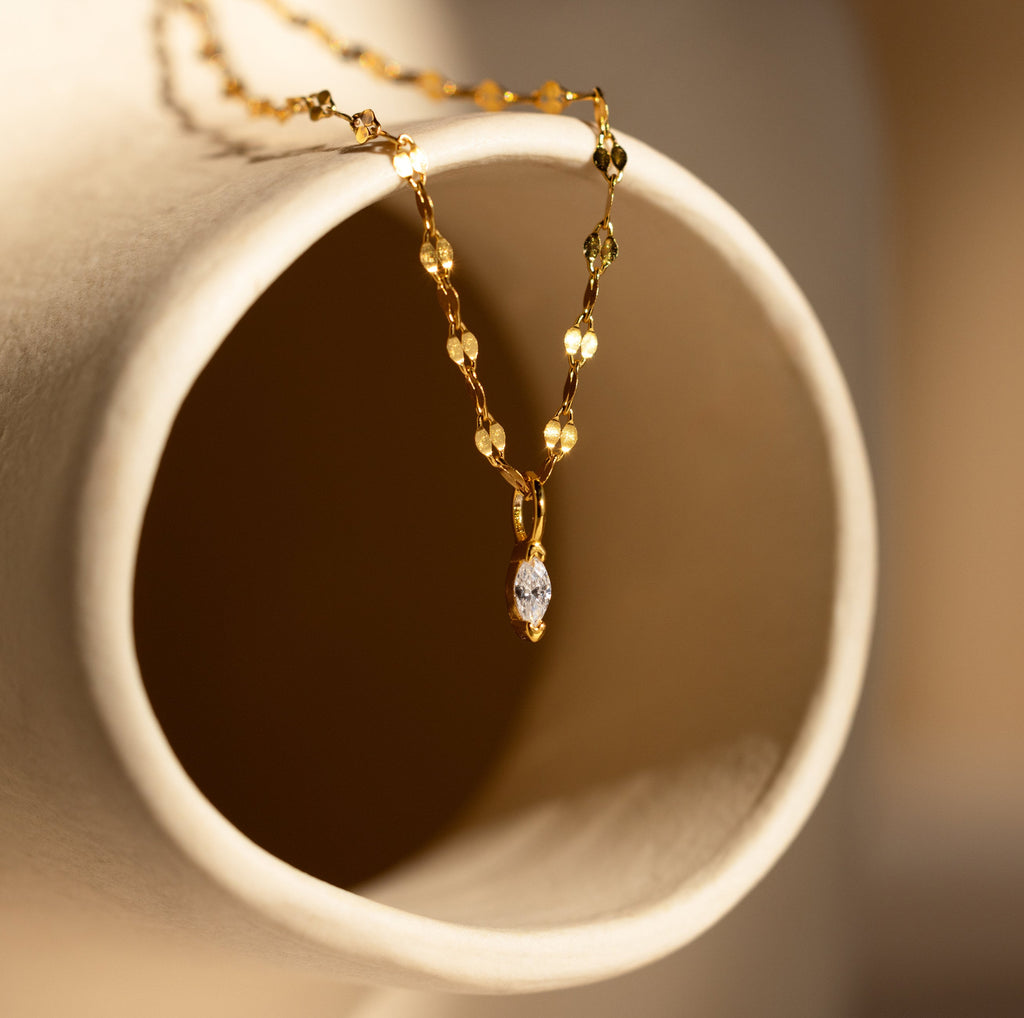 The Dainty Marquise Pendant Necklace, featuring a gold twist chain and small marquise gemstone, is draped over the edge of a cream-colored ceramic vase.