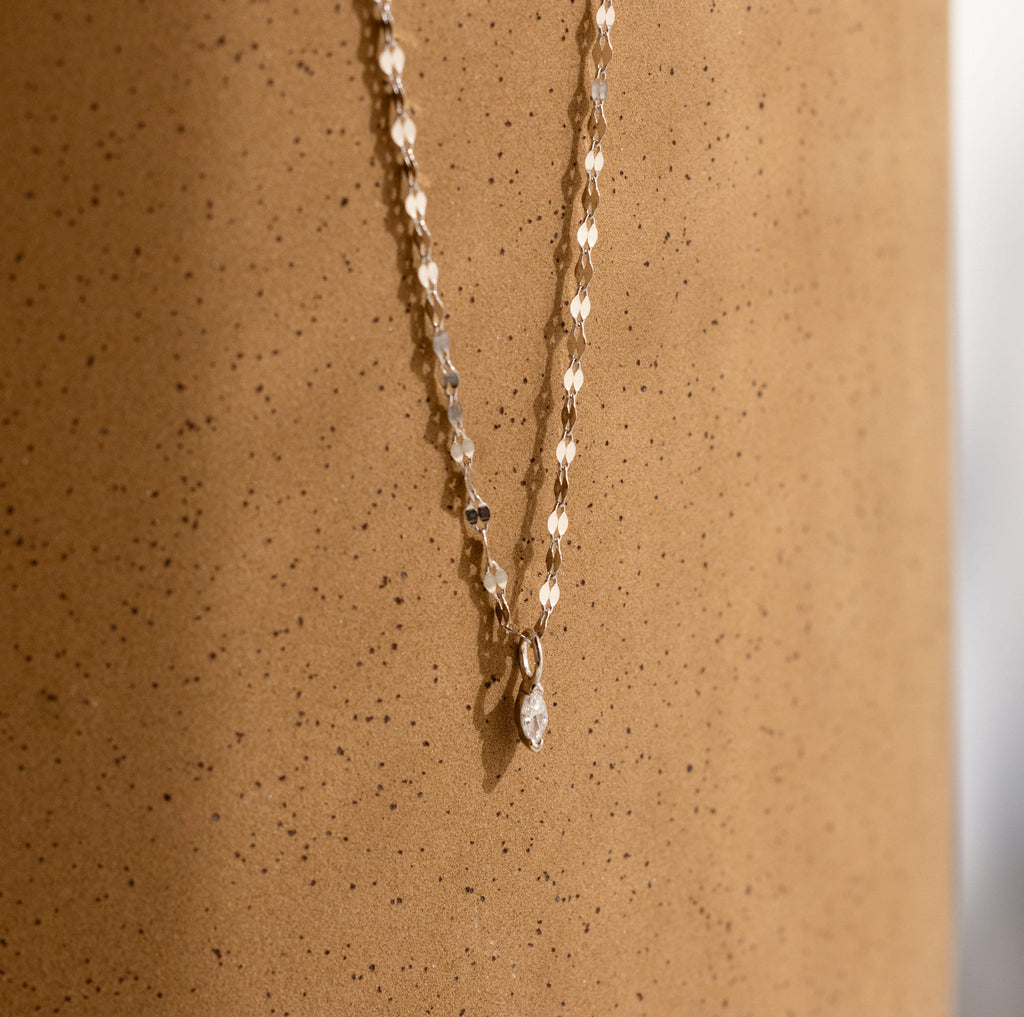 The Dainty Marquise Pendant Necklace, featuring a gold mirror twist chain and small marquise pendant, is displayed against a textured light brown surface.