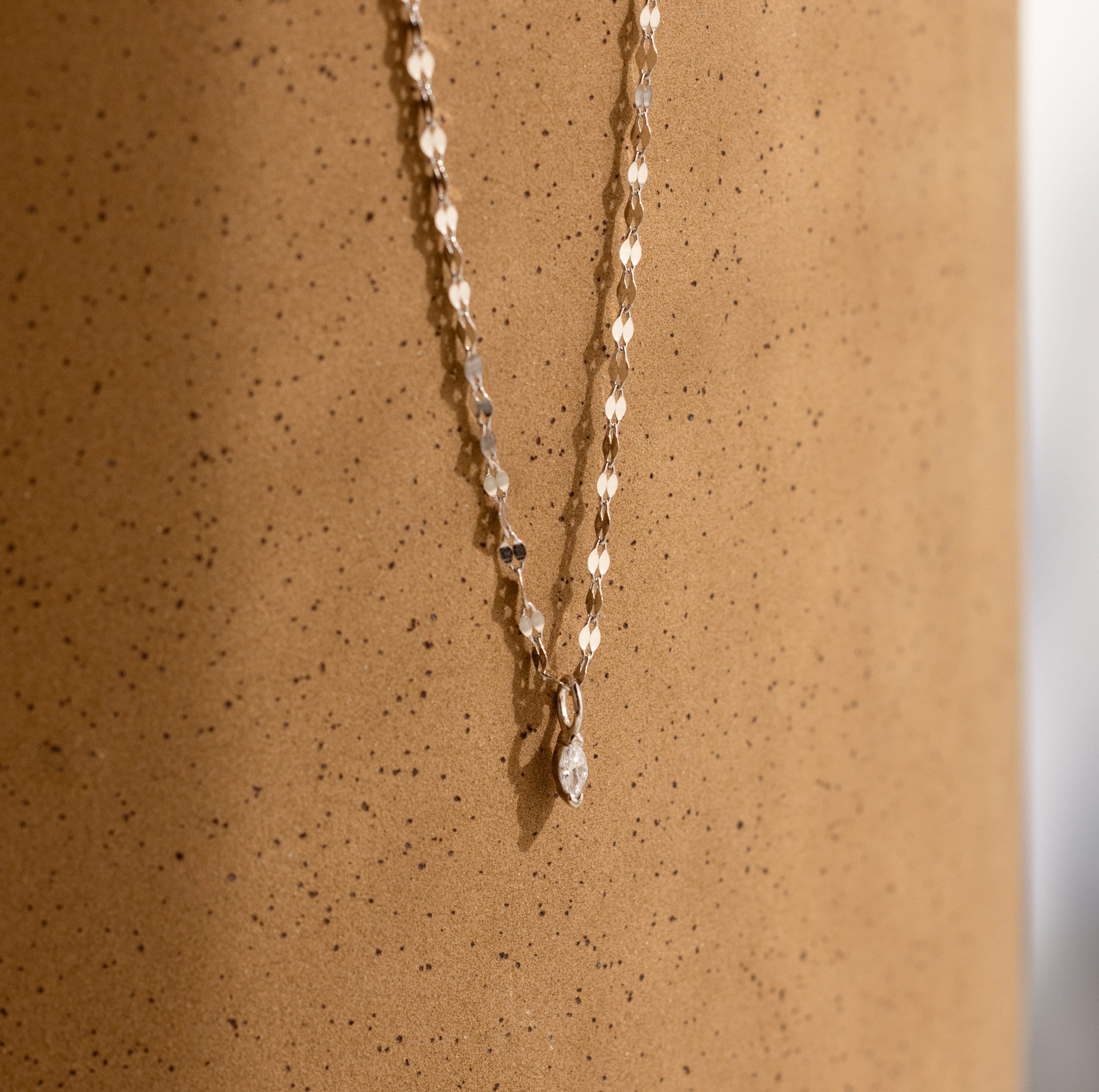 The Dainty Marquise Pendant Necklace, featuring a gold mirror twist chain and small marquise pendant, is displayed against a textured light brown surface.